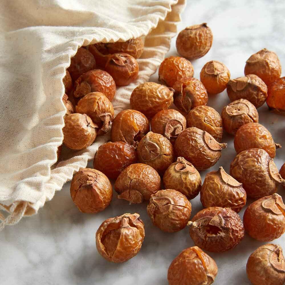 A pile of dried soap nuts spilling out of a small, white cloth bag onto a marble surface. The soap nuts are brown and appear to be dried fruit.